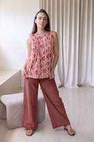 Woman wearing a floral sleeveless top with decorative pleats, high round neckline and plain pants in a room setting.