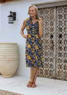 Woman wearing a sleeveless floral cotton dress with button through front, pockets and a-line skirt, standing in front of a wooden door.
