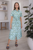 Woman standing in a room with a plant and shelves in floral printed shirt dress with detachable fabric belt, short sleeves and pockets.