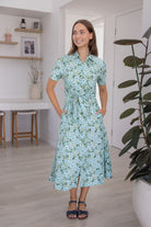 Model poses with both hand in pocket wearing a shirt style button through cotton dress with collar and a removable waist tie.