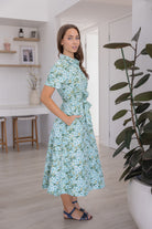 Woman wearing a 100% cotton floral dress standing in a room with a plant and shelves