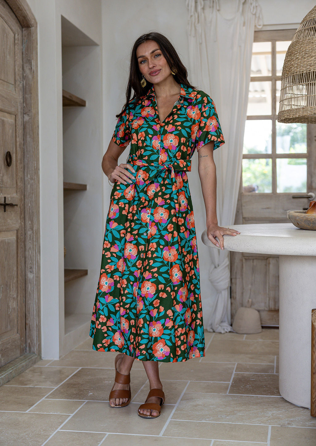 Woman wearing 100 percent cotton button through shirt dress with collar and short sleeves, in a olive green based floral print. 