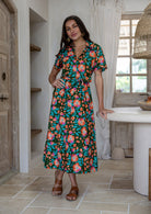 Woman wearing 100 percent cotton button through shirt dress with collar and short sleeves, in a olive green based floral print. 