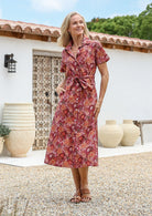 Woman wears rust pink based floral cotton shirt-style dress with collar, fabric waist tie and hidden side pockets.