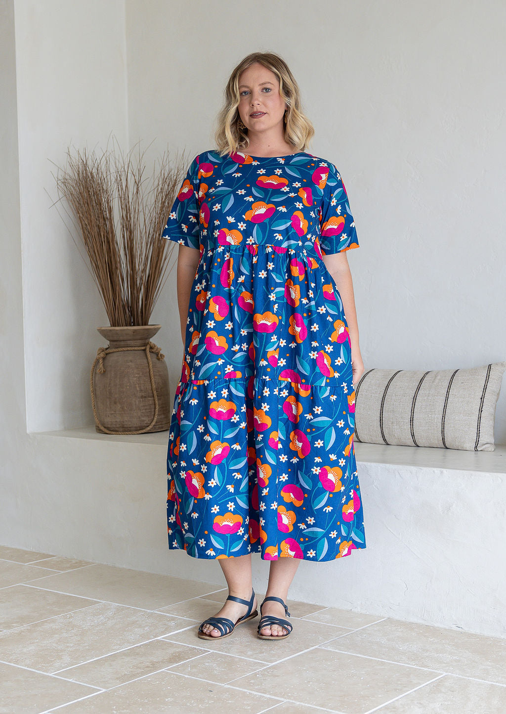 Woman wearing a 100% cotton, mid-length royal blue based dress with floral print featuring three tiered design, short sleeves, high round neckline and pockets