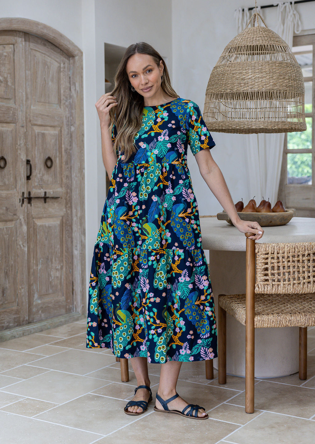 Woman wearing a 100% cotton, mid-length navy blue based dress with floral peacock print featuring three tiered design, short sleeves, high round neckline and pockets