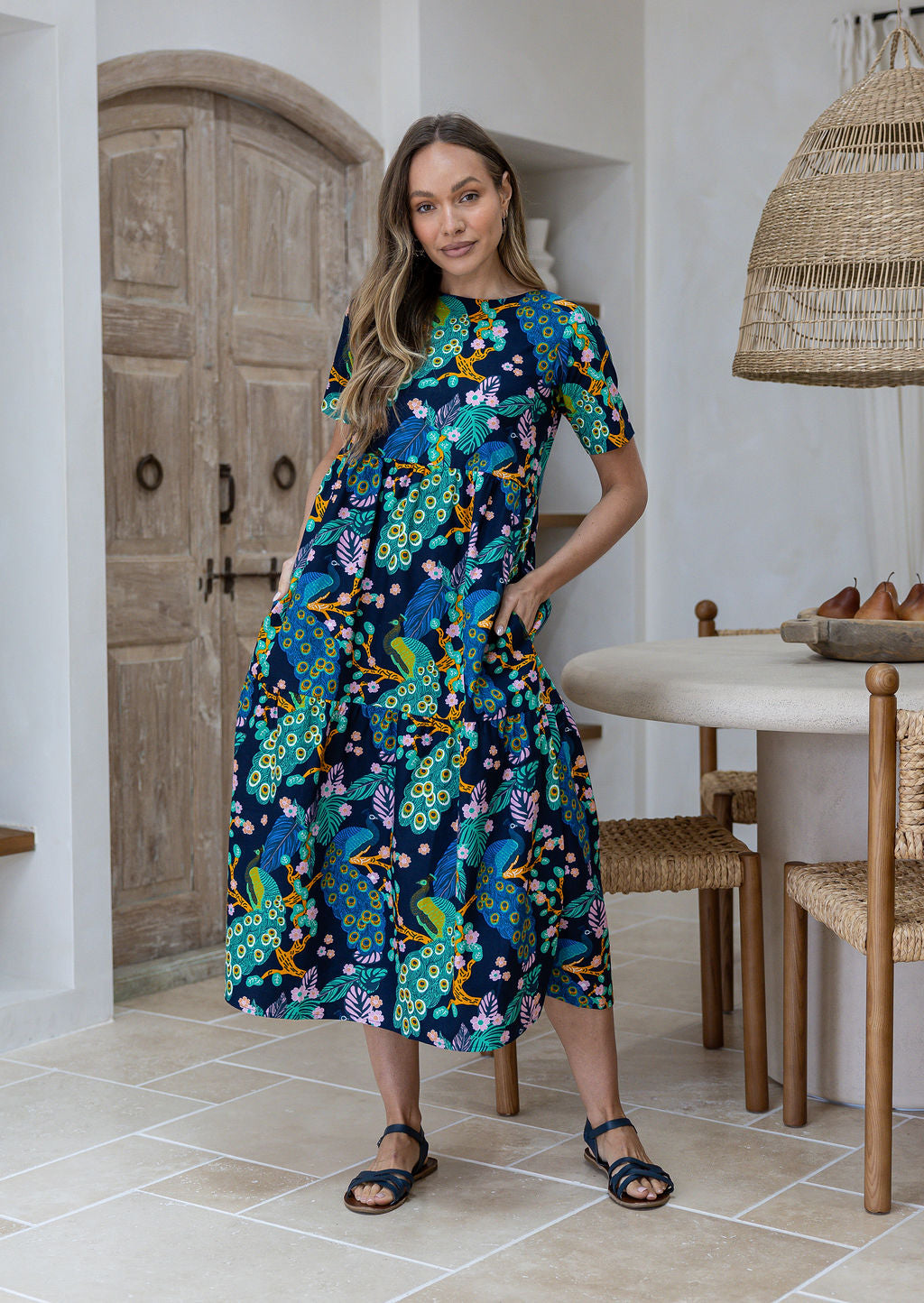 Woman standing wears a floral peacock print cotton dress with three tiered design, short sleeves and hidden side pockets