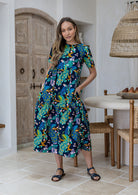 Woman standing wears a floral peacock print cotton dress with three tiered design, short sleeves and hidden side pockets