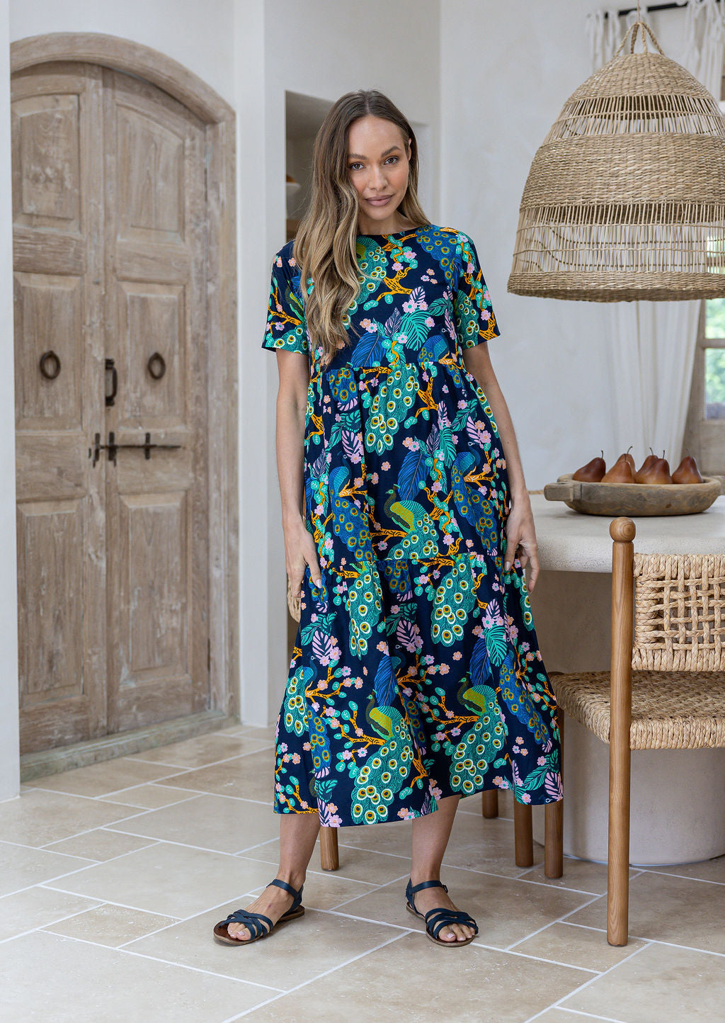 Woman is wearing a relaxed fit midi length, tiered cotton dress in navy based floral peacock print with short sleeves, round neckline and side pockets.
