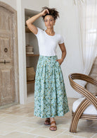 Woman wearing a white top and floral skirt standing in a room with wooden door and chair.