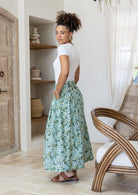 Woman stands sideways wearing a white top and sage green floral print, shin length cotton skirt with button through front and box pleats.