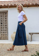 Model posing sideways wearing a 100% cotton long button through skirt in deep sea blue with pockets and box pleats 