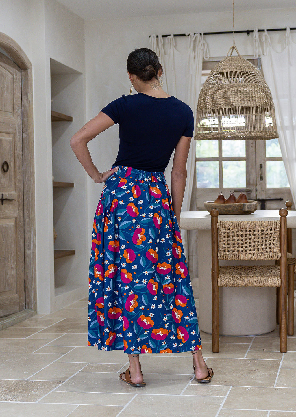 Woman stands backwards wearing a basic top and floral print, shin length cotton skirt with button through front and box pleats.