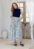 Woman stands wearing a navy top and floral print, shin length cotton skirt with button through front and box pleats.
