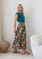 Woman stands wearing a floral print, shin length cotton skirt with button through front and box pleats.