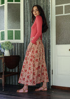 Woman stands sideways wearing a pink top and floral print, shin length cotton skirt with button through front and box pleats.