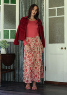 Woman in a red jacket and floral skirt standing in a room with a door and window.