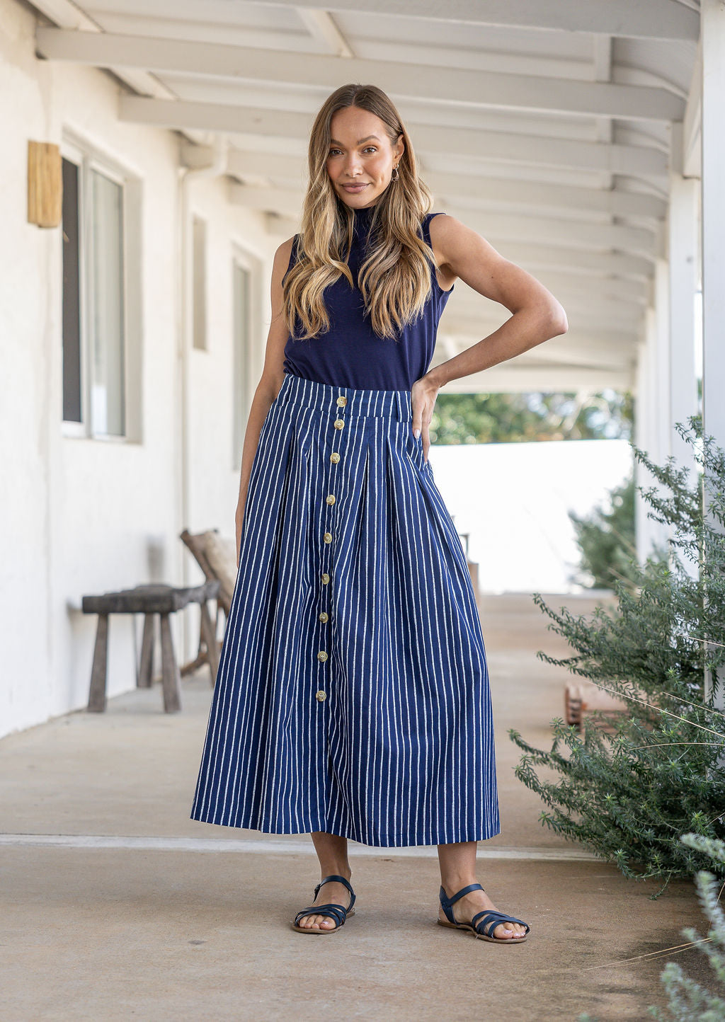Woman stands wearing a navy top and striped print, shin length cotton skirt with button through front and box pleats.