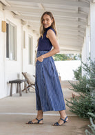 Woman stands sideways wearing a navy top and white striped on navy base print, shin length cotton skirt with button through front and box pleats.