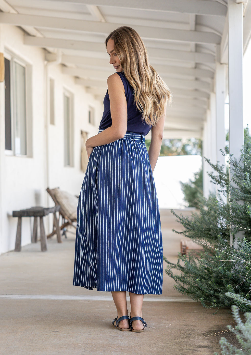 Woman stands backwards wearing a white striped on navy base print, shin length cotton skirt with button through front and box pleats.