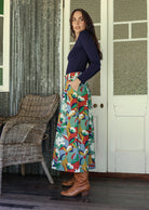 Woman stands sideways wearing a floral print, shin length cotton skirt with button through front and box pleats.