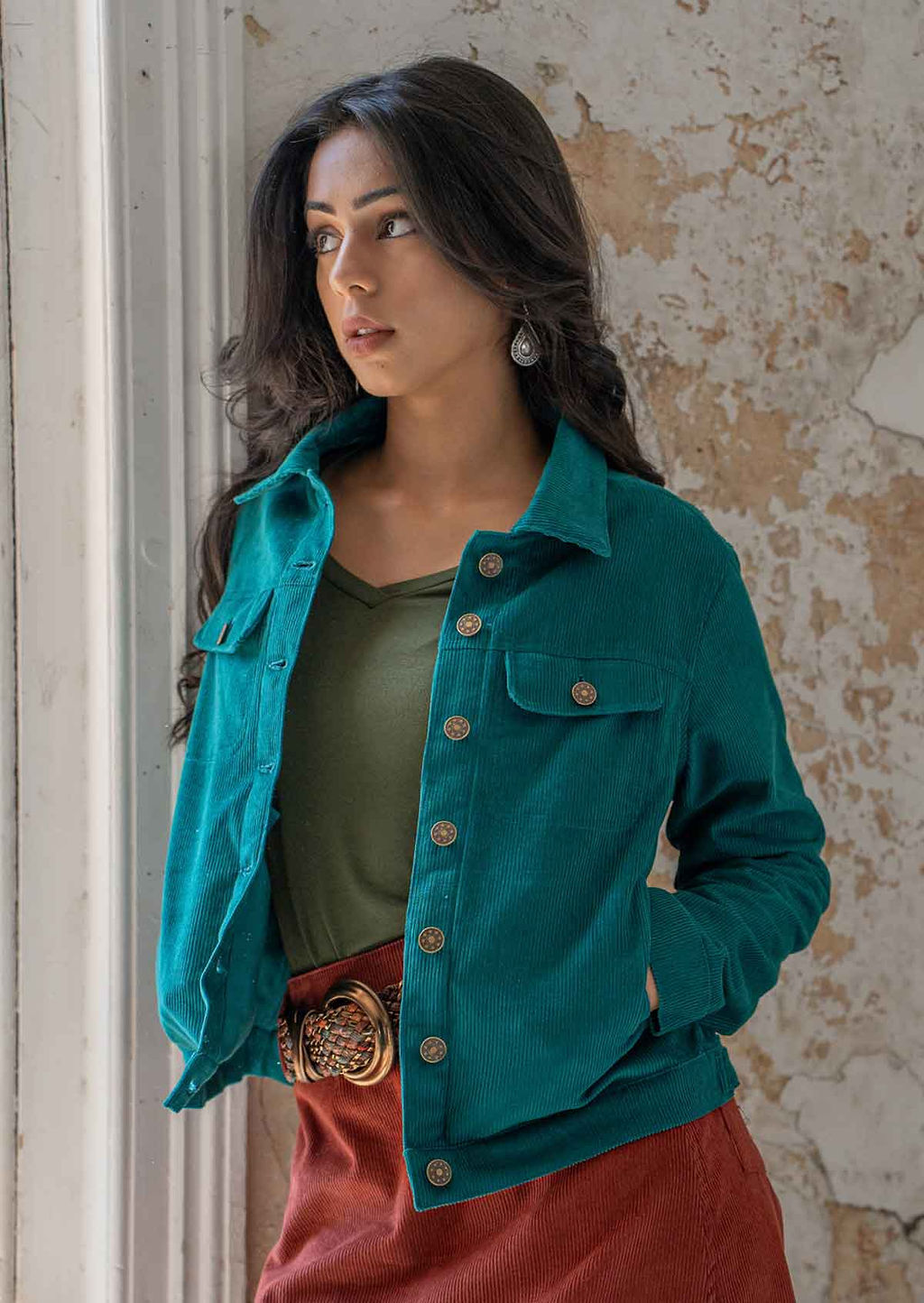 Woman wearing teal corduroy jacket with breast and front pockets, and brass buttons with hands in pockets 