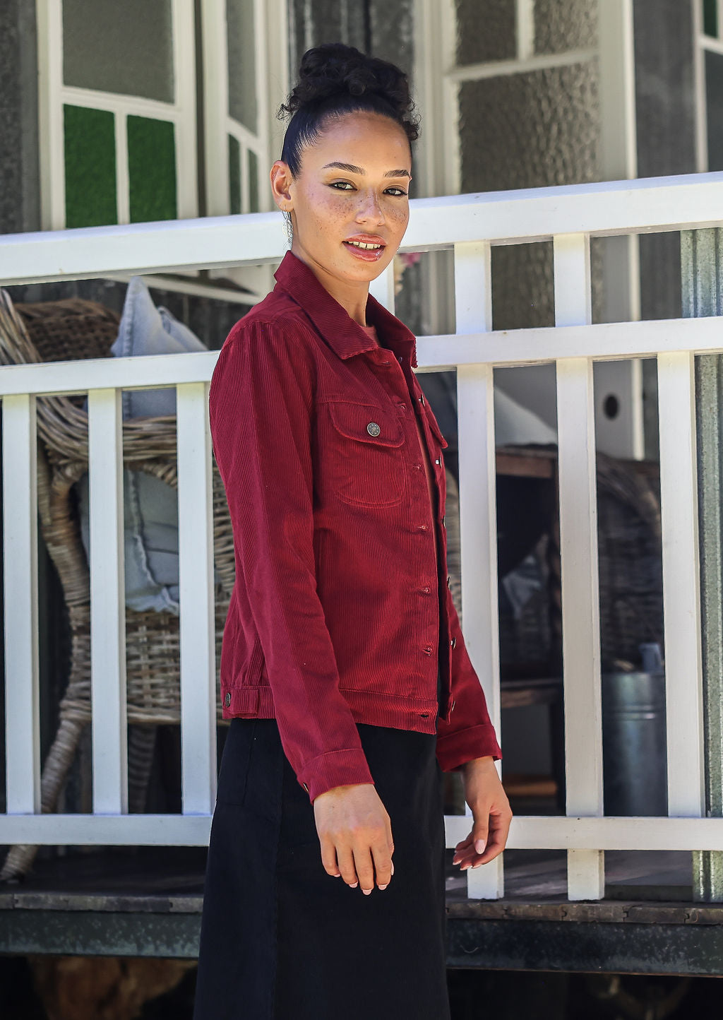 Woman stands sideways wearing a corduroy jacket and black corduroy skirt against a white railing.