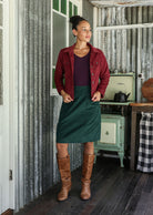 Woman in a deep, luxurious, and intense red-purple hue jacket and green corduroy skirt standing in a rustic room with a stove and checkered tablecloth.