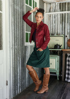 Woman in a corduroy jacket and green skirt standing in a rustic kitchen.