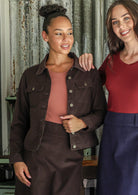 Two women standing together, on wearing a brown corduroy jacket and skirt combo, the other in a red top and blue corduroy skirt, against a rustic metal wall.