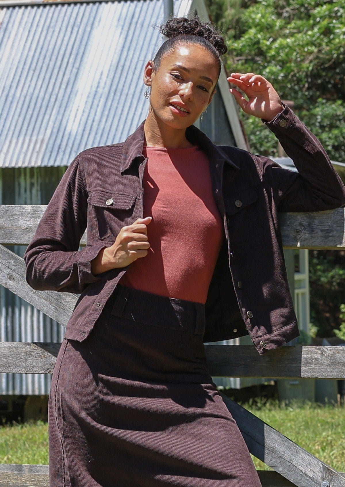 Woman wearing a brown cotton corduroy jacket and skirt, paired with a pink top, in front of a wooden fence