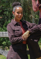 Woman in a brown corduroy jacket and pink top outdoors with greenery in the background