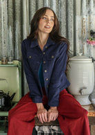 Woman in a navy blue jacket and pants sitting on a wooden stool in a rustic kitchen.