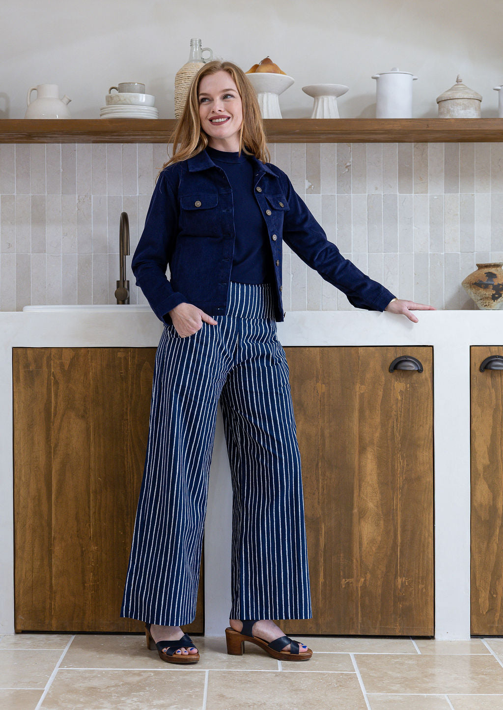 Woman in a kitchen wearing a navy jacket and striped pants.