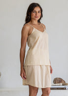 Woman wears lightweight cotton cami in nude tone, with V-neckline, spaghetti straps and a picot trim, paired with a matching cotton slip skirt in nude