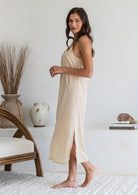 Woman stands side on wearing nude lightweight cotton maxi slip with V-neckline, bust darts, side splits and picot trim hem