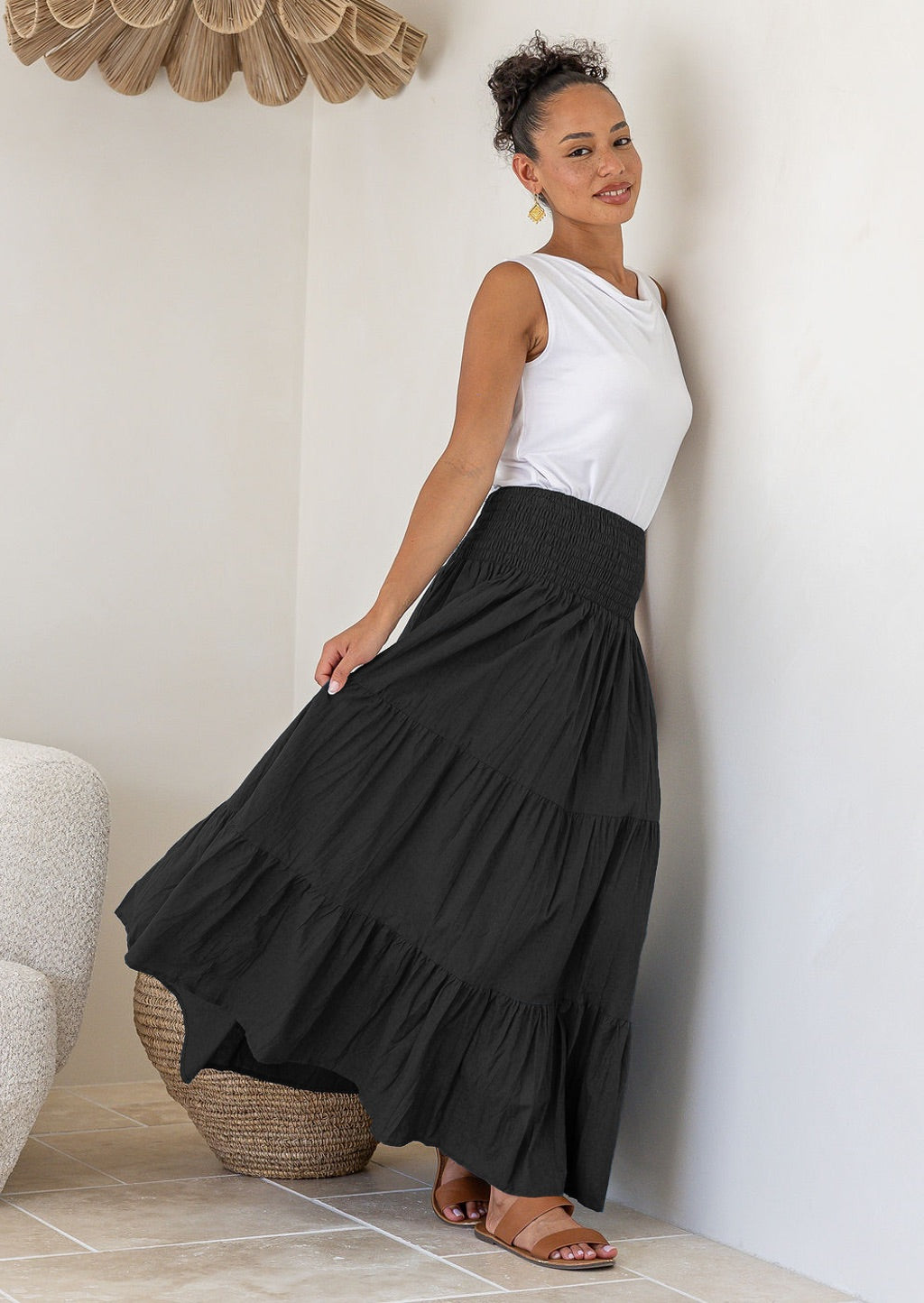 Woman leaning on wall wearing a white singlet top and black cotton tiered maxi skirt with wide ruched waistband, holding skirt out to the side