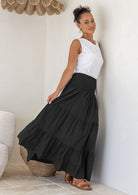 Woman leaning on wall wearing a white singlet top and black cotton tiered maxi skirt with wide ruched waistband, holding skirt out to the side