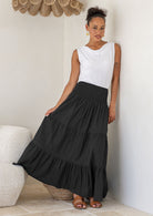 Woman wearing a white singlet top and black cotton tiered maxi skirt with wide ruched waistband, holding skirt to side
