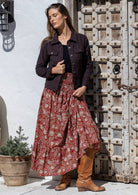 Woman poses in earthy red based floral cotton maxi skirt paired with a dark brown top, a dark brown corduroy jacket and tan boots