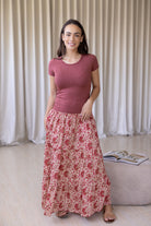 Woman wearing a pink top and cream based floral skirt standing in a room with a curtain background.