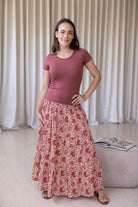 Woman wearing a pink top and floral skirt standing indoors with a curtain background
