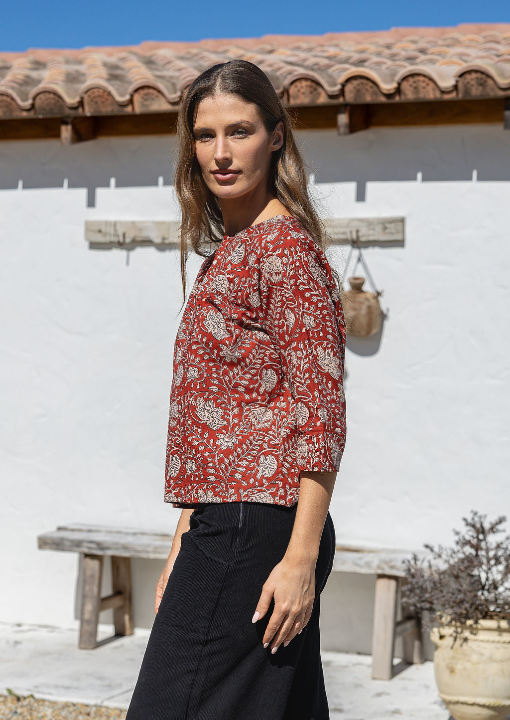 Woman stands side on in an earthy red based floral cotton top with 3/4 sleeves