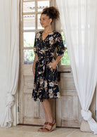Woman wearing over the knee length wrap dress in black based cotton print and a hand in pocket.