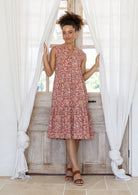 Woman wearing a rose pink based floral sleeveless, knee length dress with a buttoned bodice, tiered skirt and pockets. 