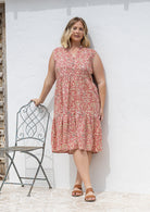 Woman wearing a knee length cotton sleeveless dress with buttoned bodice and  two tiered skirt in rose pink based floral print.