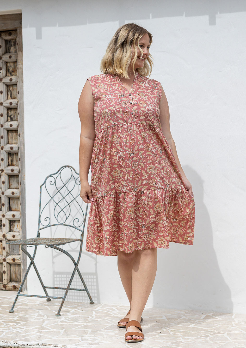 Woman wearing a rose pink based floral sleeveless, knee length dress with a buttoned bodice, tiered skirt and pockets. 