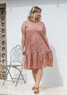 Woman wearing a rose pink based floral sleeveless, knee length dress with a buttoned bodice, tiered skirt and pockets. 