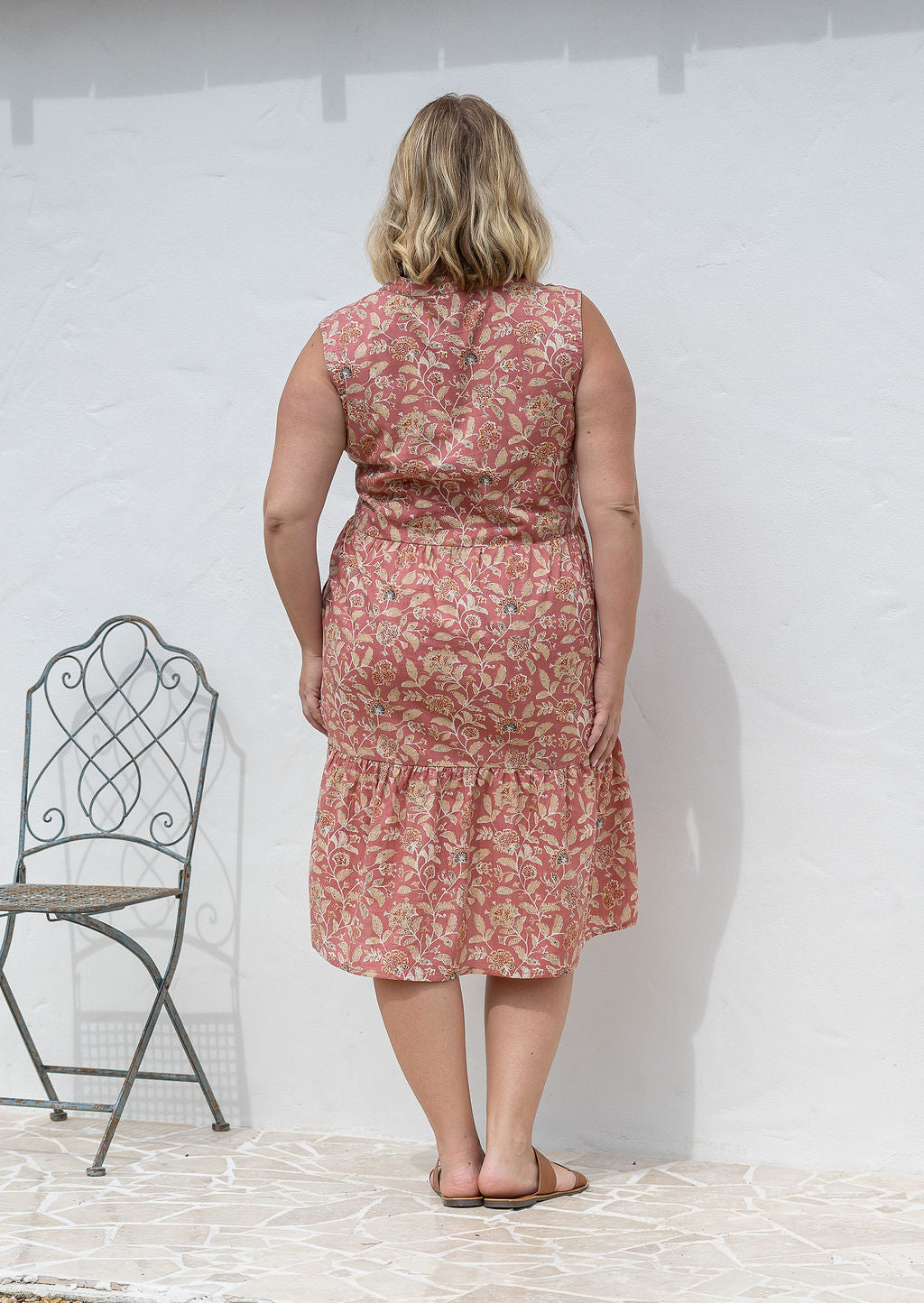 Woman stands backwards wearing a sleeveless rose pink floral cotton dress with pockets, mandarin collar and a two tiered skirt.