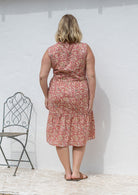 Woman stands backwards wearing a sleeveless rose pink floral cotton dress with pockets, mandarin collar and a two tiered skirt.
